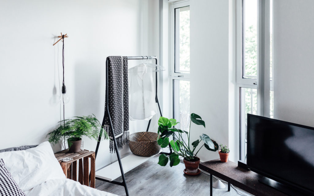 A crisp and clean white painted bedroom. There are a few plants, a tv, a clothing rack, a crate as a night stand and a bed with a white comforter. 