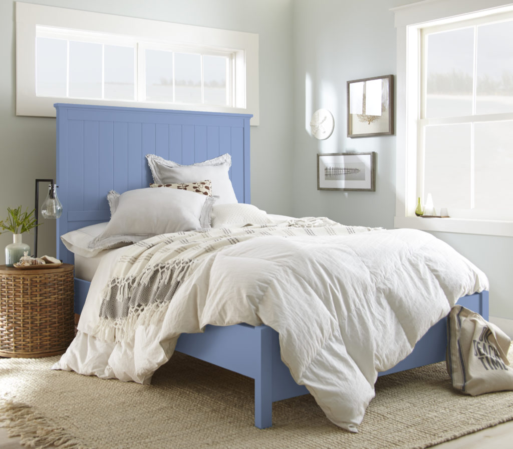 A well-lit and airy coastal style bedroom with a blue wooden bedframe and fluffy light neutral bedding. 