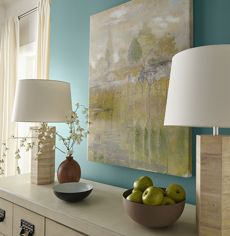 A living room accent wall painted in fresh blue color called Dragonfly. A dresser top decorated with two stylish lamps, a two simple bowls and a round vase with white flowers. 