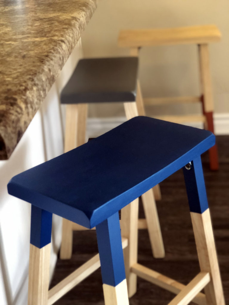 This image shows three painted bar stools, in the foreground is a partially painted stool in Nocturne Blue with the seat painted and a painted portion almost halfway down the legs. 
In the background is a stool partially painted in the color Shadows only the seat is painted. Lastly, in the background of this image is a blurred stool with the legs partially painted in Kalahari Sunset. 