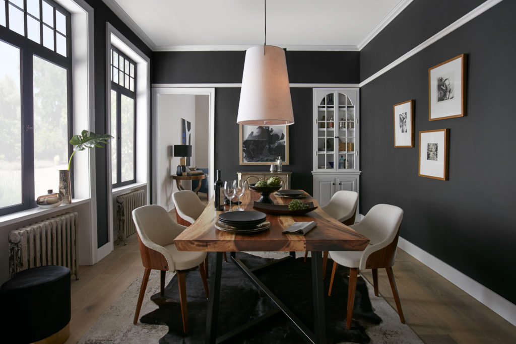 A stunning black dining room with room with walls painted in color called Broadway.   Off White moldings, natural wood dining table and light upholstered wooden chairs. 