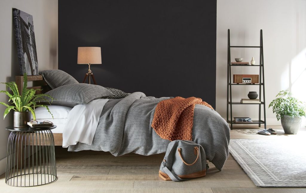 A large white  modern bedroom with a black accent wall,  the bed is dressed in cozy, soft layers of white, gray, and rusty orange linens. 