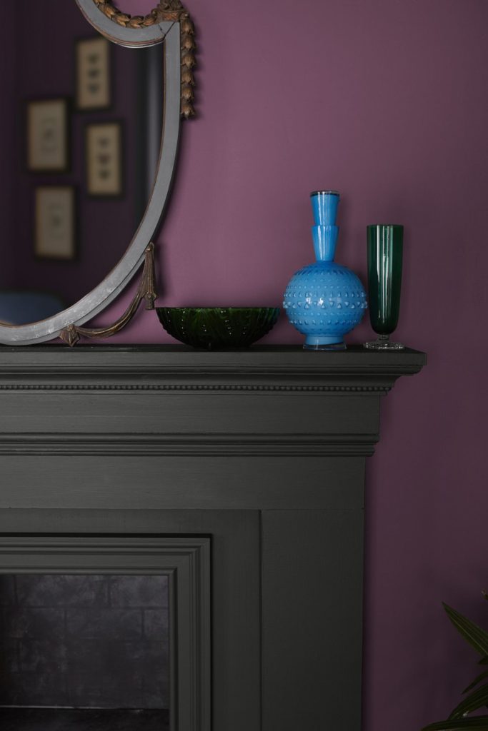 – A close-up detail of a living room wall and fireplace, the wall is painted in a bold and saturated color called  Euphoric Magenta. The fireplace is painted in a black color called Broadway. 