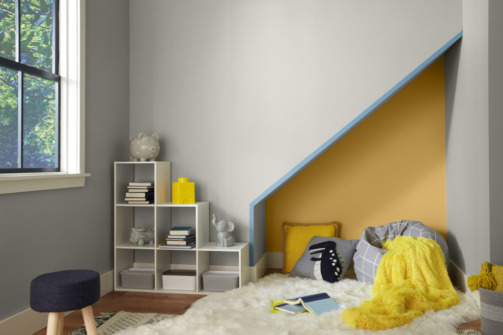 A modern cozy nook under the stairs.  The room is meant to be used as a kids play, sleep and relax area.  The main color of the wall is a light gray and the accent color are a bright yellow and a pastel blue. The decor elements include fluffy pillows and blankets.  