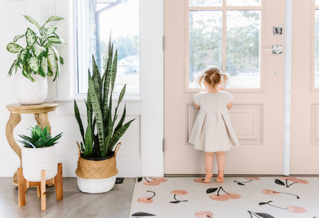 A home entry with a pink double door. 
 There is a little girl looking out the door. The entry is decorated with pink cherry pattern rug and plants placed in modern planters and wooden stands. 