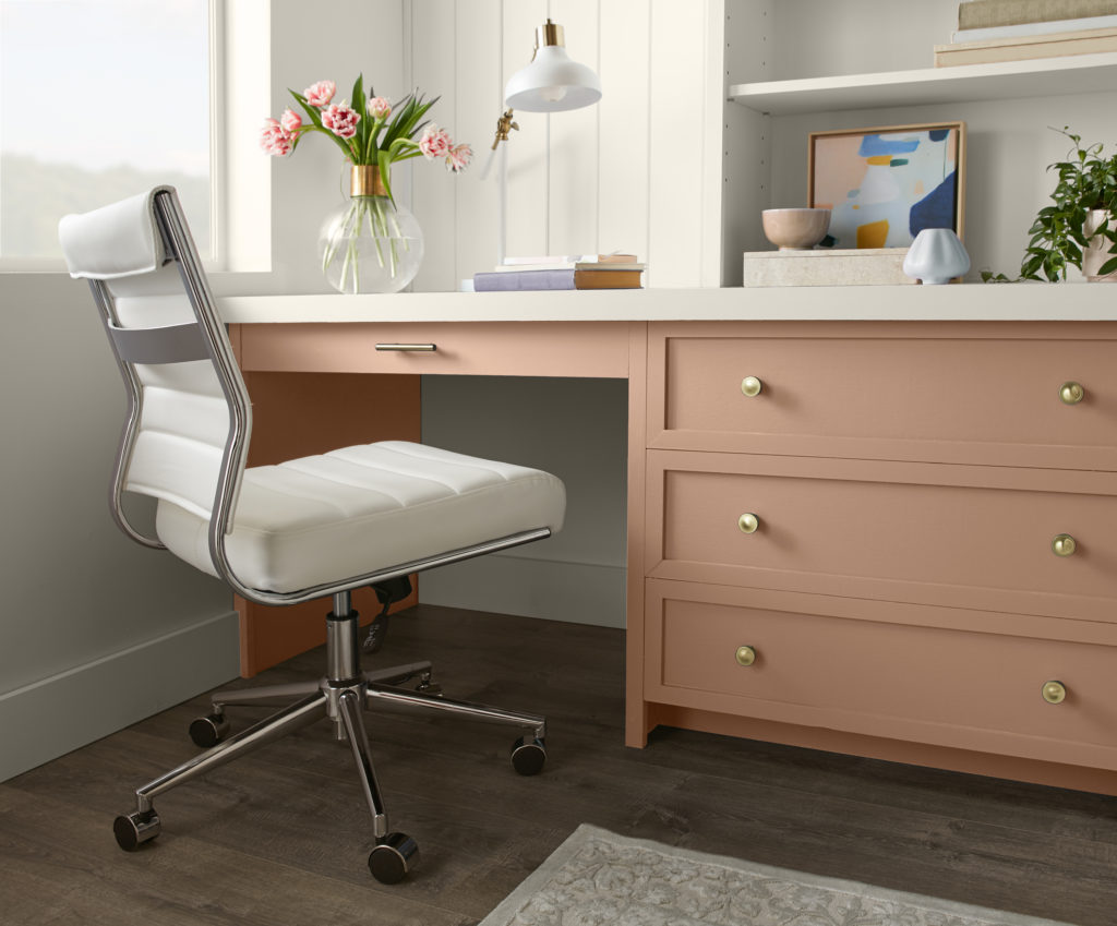 A built-in desk area in the corner of a room, featuring a terra cotta desk and bookshelves.  The walls on the room are white.  An adjustable white leather chair and some colorful accessories make up the rest of the office space. 