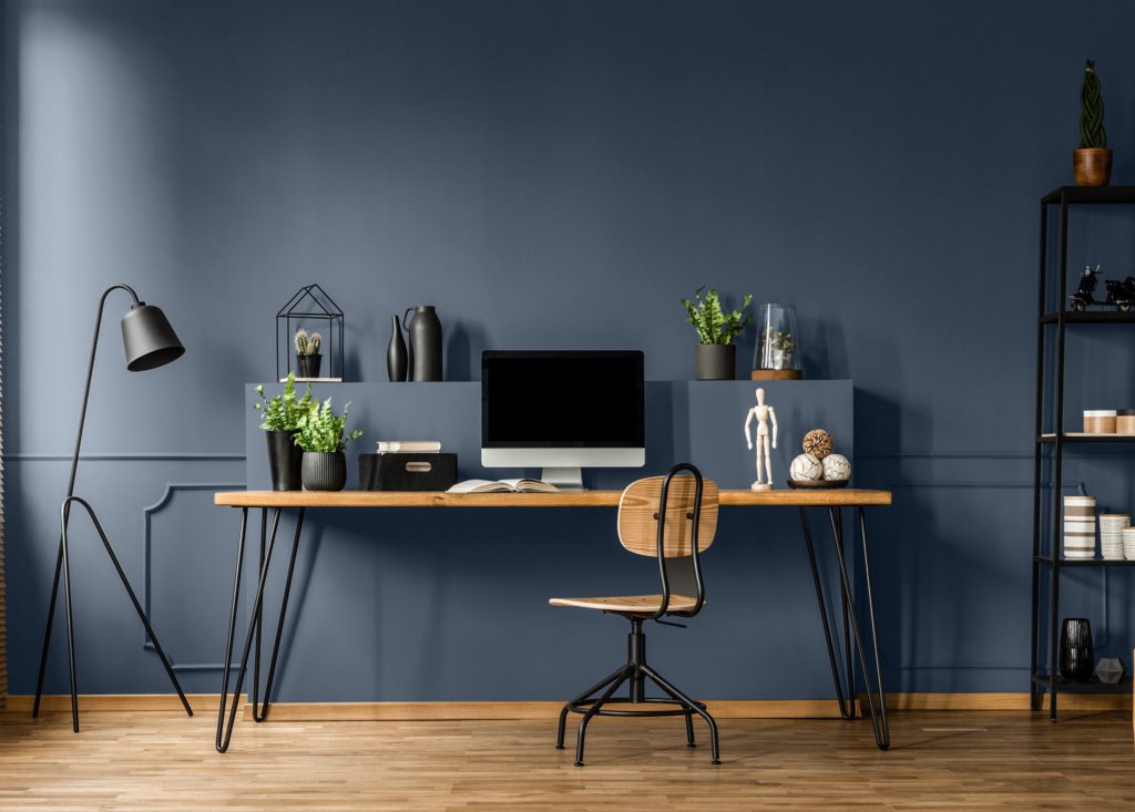 Chair at wooden table with computer monitor and plants in dark blue and  spacious home office interior.  