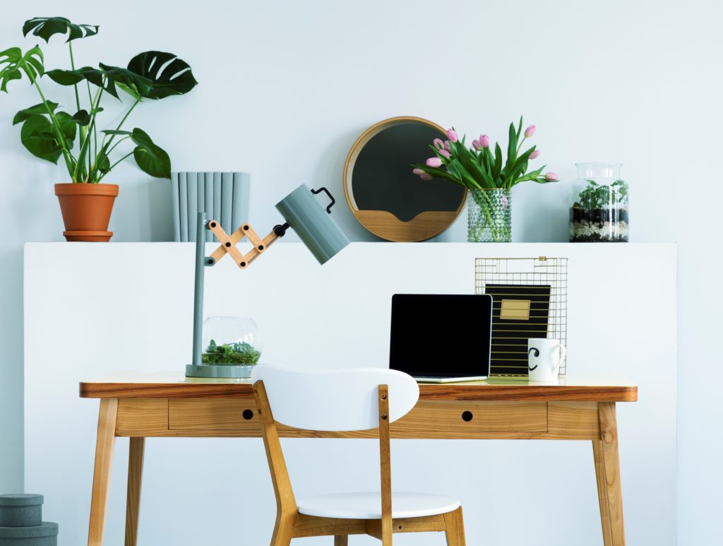 A wall shelf with flowers and fresh plant, decor and books in white living room interior with study corner desk with lamp and mockup laptop.  The color in the wall is a light aqua called Beach Foam. 