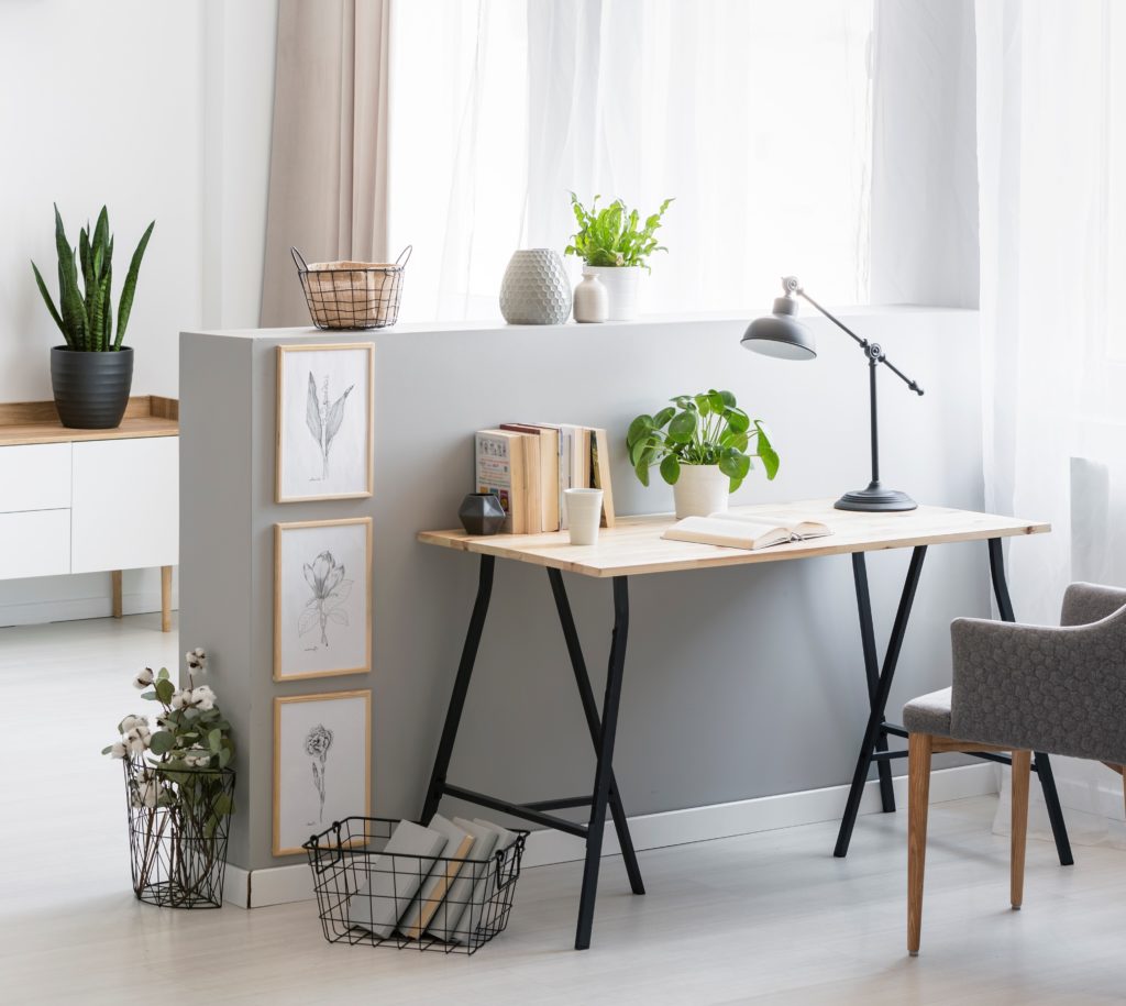 A short wall painted in a light gray paint color. A simple yet stylish home office area with a gray armchair at desk with plant and lamp.  