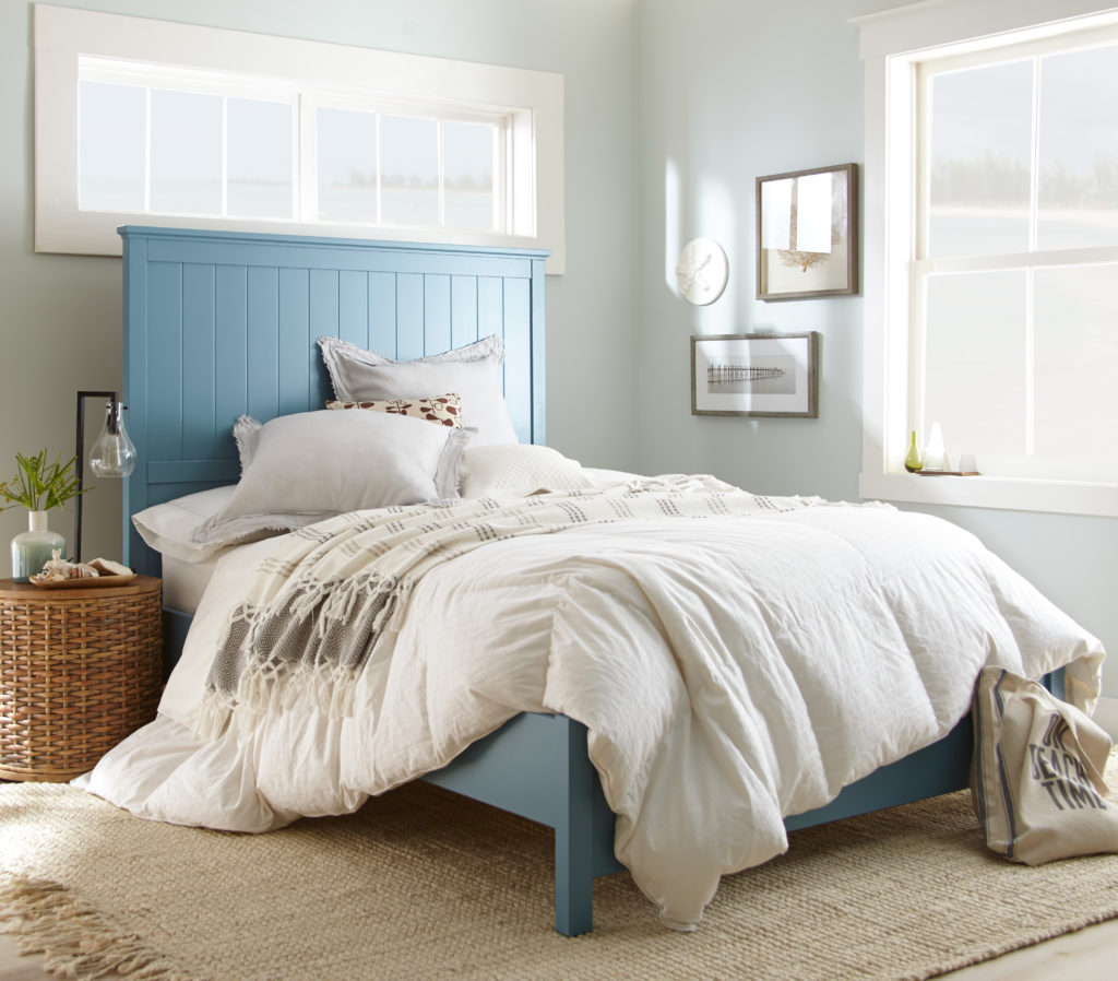 A cottage style bedroom, painted in light and airy light green color. 
The wooden bedframe is paint in a blue paint color. There is a lot of natural light coming into the room as there are two large windows allowing the light to com it. The decor is beach cottage chic style. 