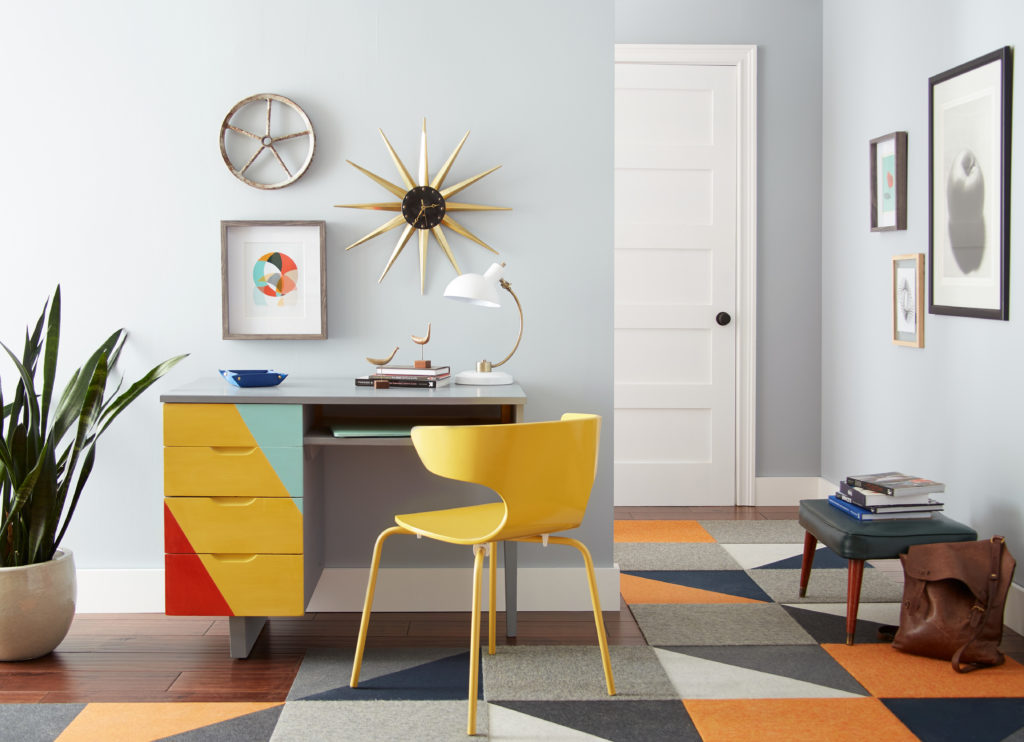 A Mid Century Modern homo office space placed next to a hallway.  The composition of the door includes a desk with a colorful geometric pattern painted on the front and a bright yellow chair.  The color on the walls is called Meteor Shower. 