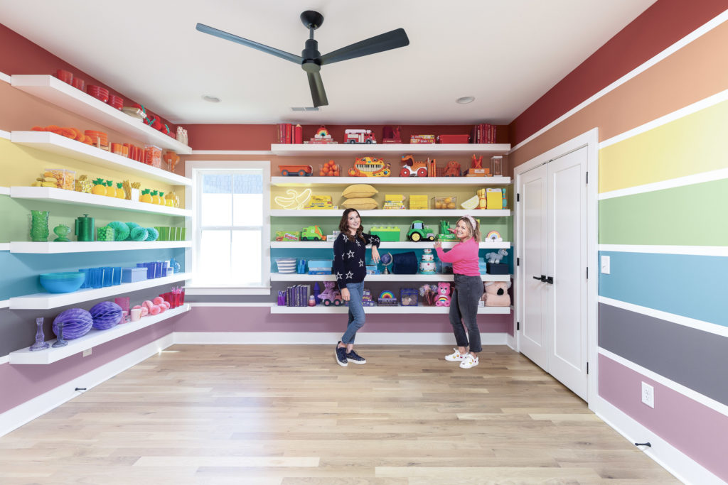 A room painted with a whimsical and colorful rainbow  in a vertical direction in all four walls of the room.  There are organizational open and free standing white shelves in two walls.  Clea and Joanna are standing intron of one set of shelves. 