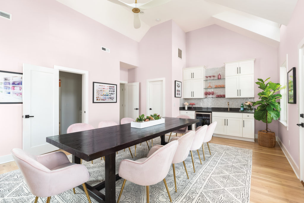 A large dining set and kitchenette room which serves as the teamwork and collaboration room. The room is painted in a light pink color called Moxie and the the ceiling is high and white with a white fan. 