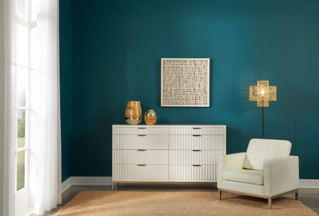 A one side angle of a bedroom, the wall being being featured is a deep ocean inspired teal called Ocean Abyss.  There is light color dresser and upholstered chair in front of the wall which creates a nice contrast between the wall and the furniture. 