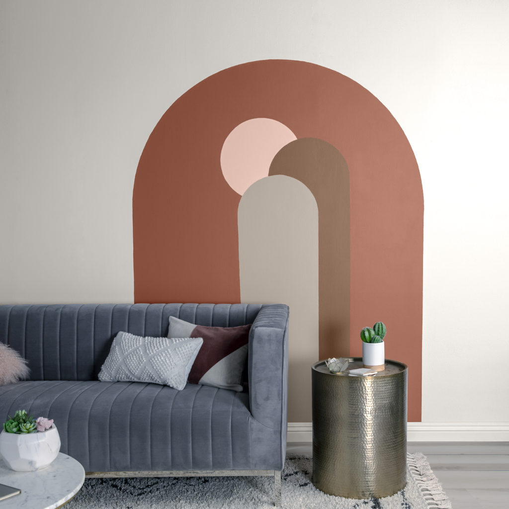 An urban boho living room featuring a painted arches mural.  There is a large arch, two smaller arches inside the large arch and a circular shape emulating a sun.  Wall mural wall is painted in a white color called Whisper White.  The arches are painted in a warm autumn inspired color palette. The living room includes a gray velvet sofa, a hammered brass side table and some fun decorative décor elements including a succulent plant and a cactus plant. 