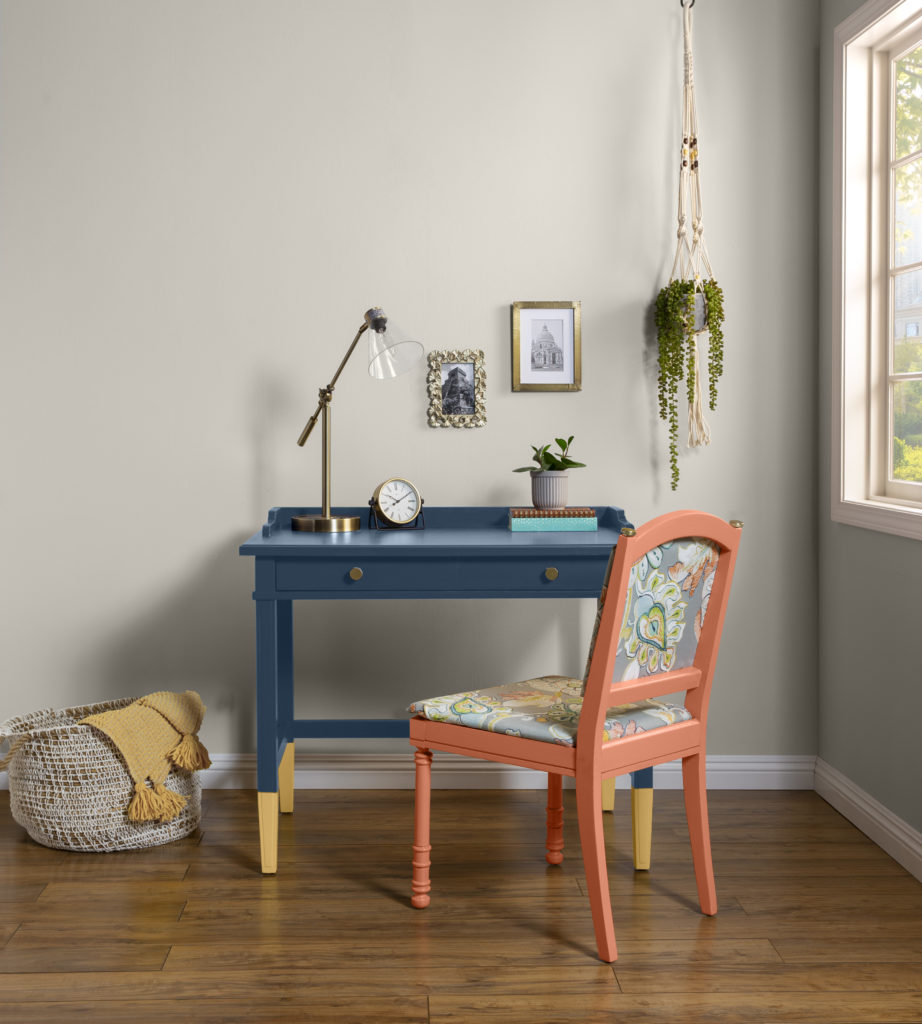 A home office with walls painted in a gray hue. The desk is a deep bold blue color with the legs in a vibrant yellow hue. The chair of the desk is a muted coral with a floral-patterned upholstery.