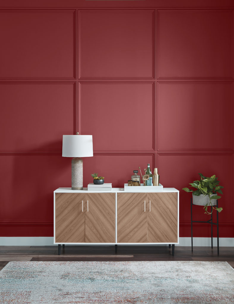 The vertical angle of a dining room, this angle includes a sideboard or buffet, also known as a buffet table decorated with a lamp, books, and a serving tray full with bar items.  
The wall is painted in a red color called Lingonberry Punch.