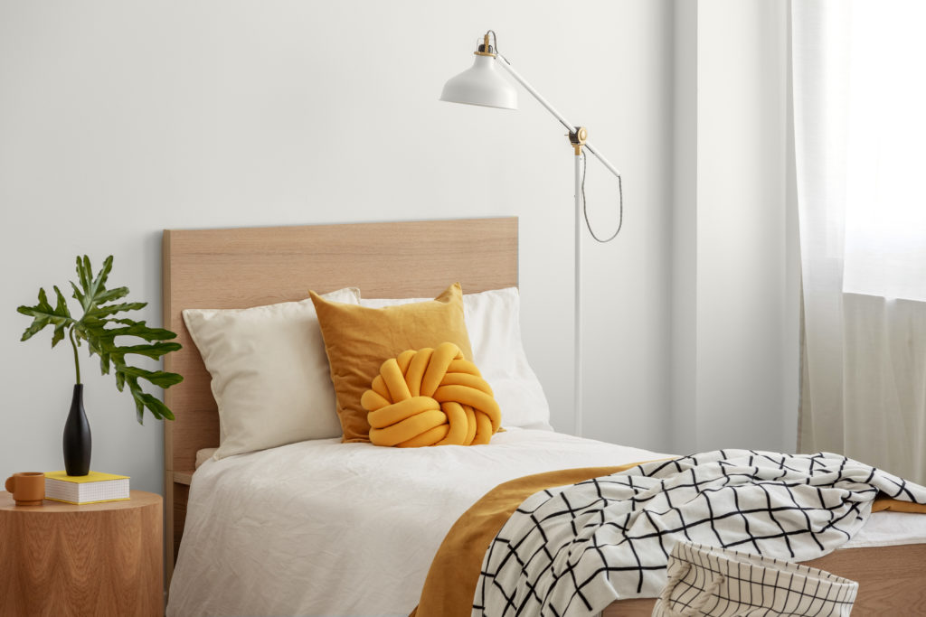 Yellow pillows and cozy blanket on single bed in elegant hotel room, green leaf in black vase on wooden bedside table. 