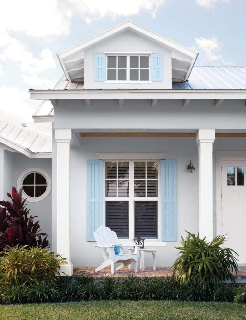 A beach cottage painted in a light blue color, the trim is white and there is an Adirondack chair on the front porch. 