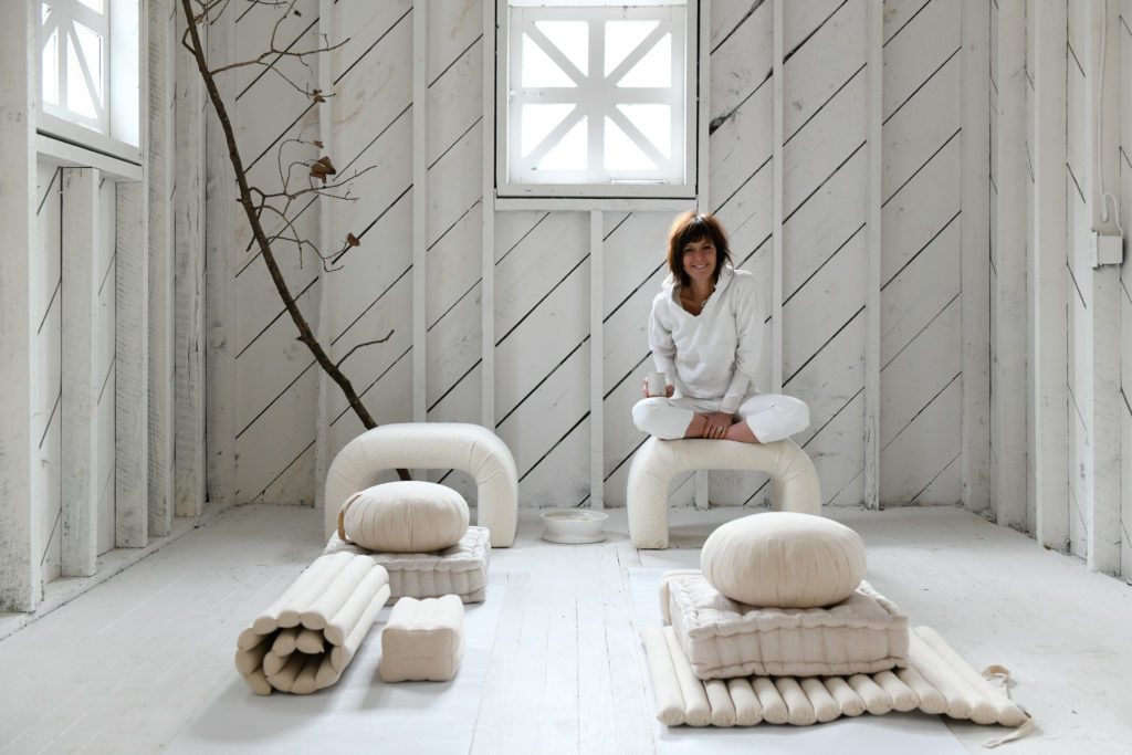 A person sitting on a white u-shaped chair in a newly renovated yoga room that is painted in all white with all white furnishings.