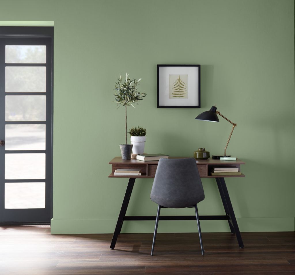 A casual office area carved in a living room setting, there is a modern desk and comfy chair, to complete the look, there are some casual desktop decorative elements. The wall color is a mid-tone green called Laurel Tree. 