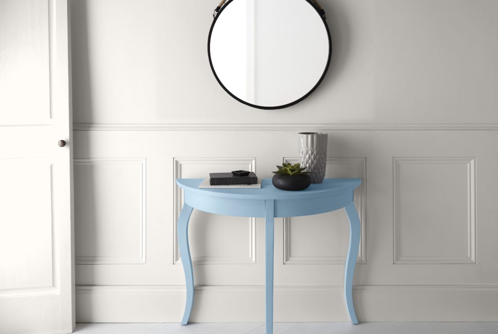 An all white hallway with a small blue console table.  There is a simple round mirror and decor items on top of the table. 
