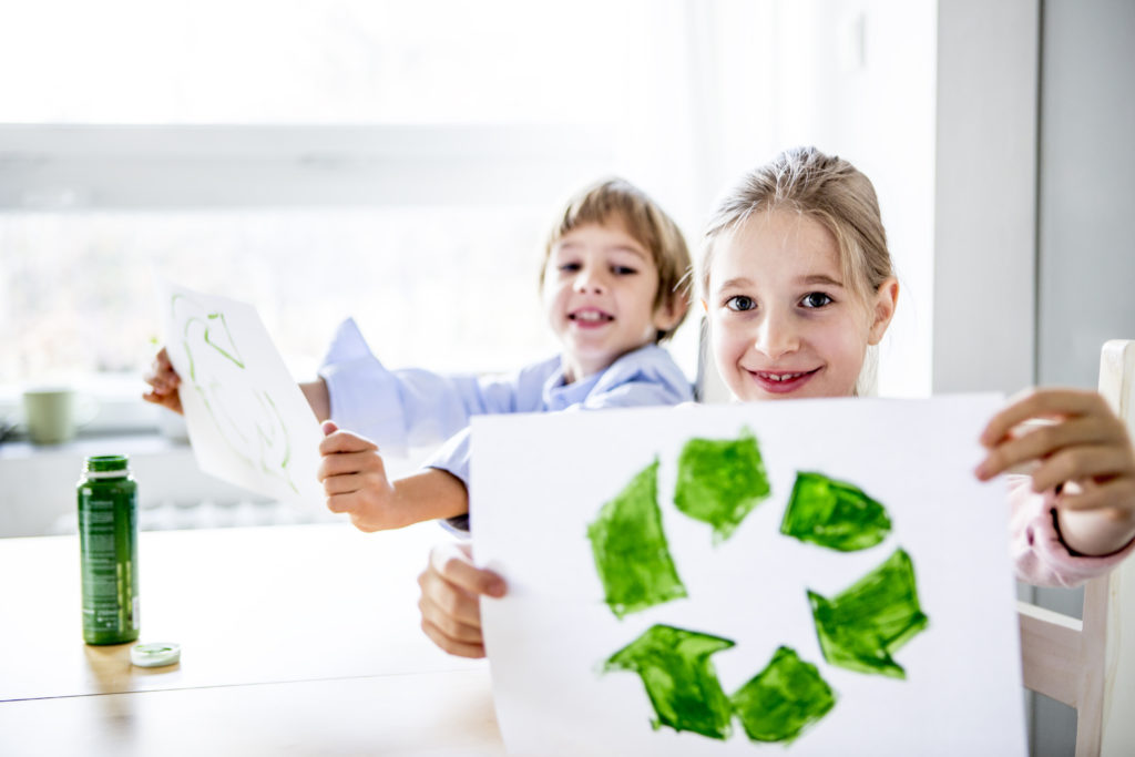 Two young children painting. One of them is holding a piece of paper showing a painted recycle sign; green arrows going in a circle.