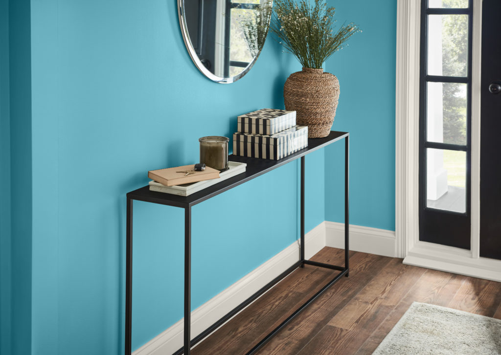 A contemporary home entry, the walls are painted in a blue color called Explorer Blue. There is a metal accent table an oval mirror mounted on the wall.  