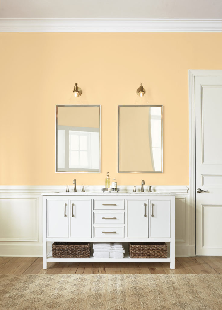 A traditional style bathroom painted in a mid-tone yellow that feels cozy and relaxing. The room features a double sink vanity and  beautiful woodwork on the lower wall area.  