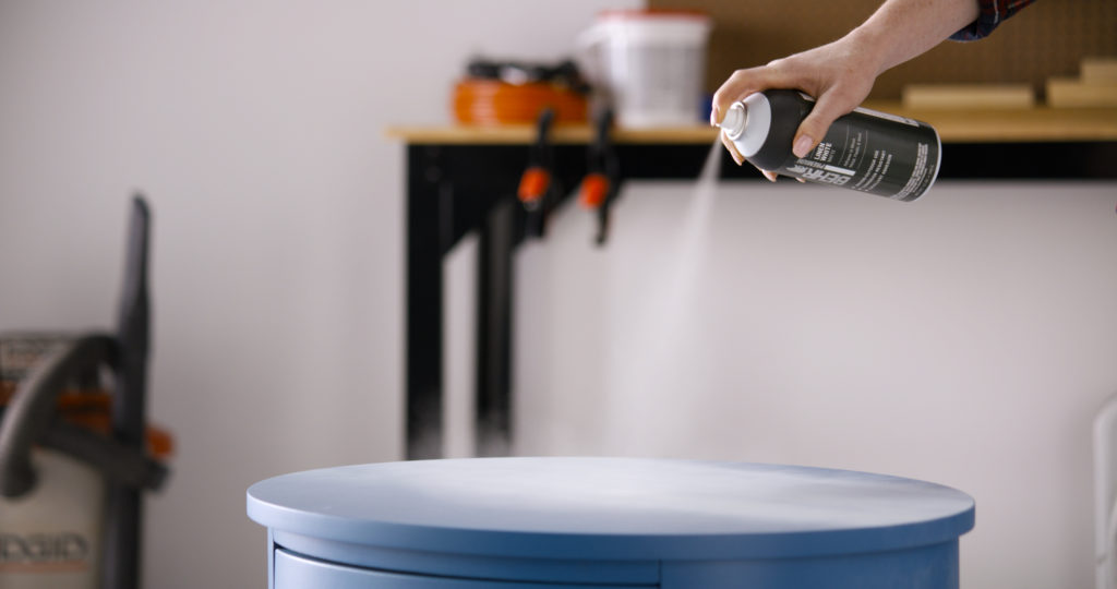 A person spraying the top of a nightstand with a white spray paint.