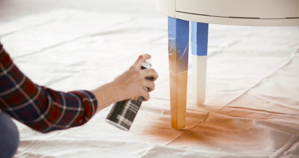 A person spraypainting the bottoms of a white nightstand with a gold color spray paint.