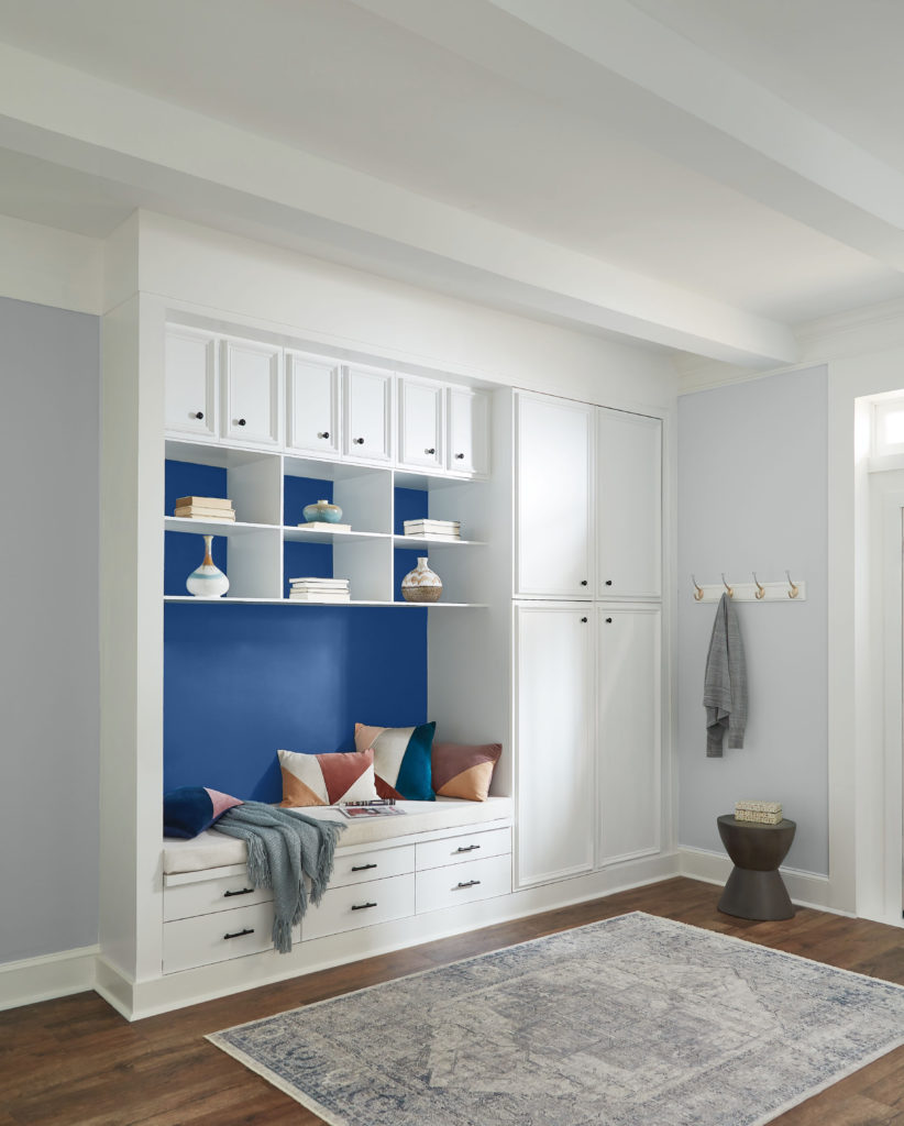 A modern mudroom/entry area painted in light gray color, white trim and cabinetry. There is a dark blue accent color being featured that highlight the built-in sitting area. 