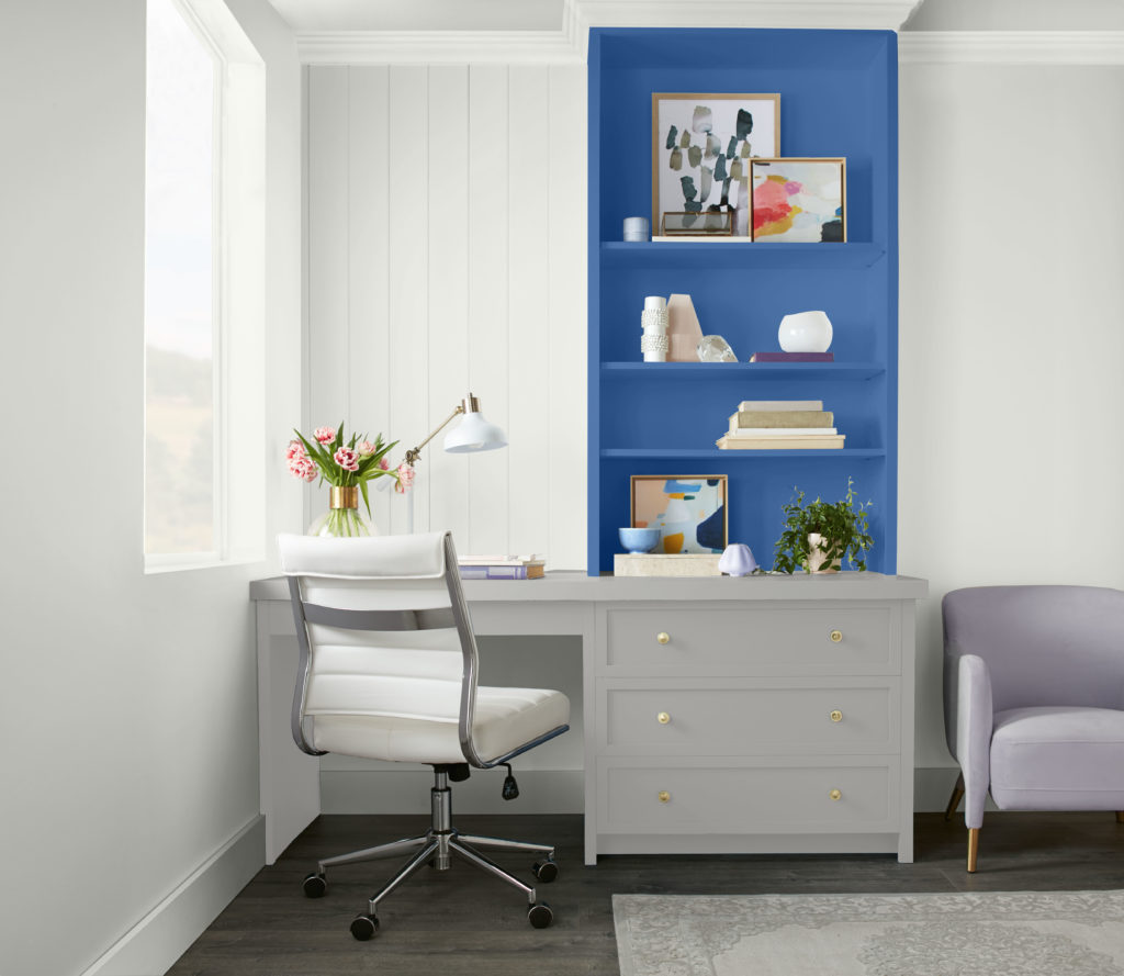 A casual home office featuring a built- in desk,  a bright blue color serves a fun background to the shelving area and décor elements. 