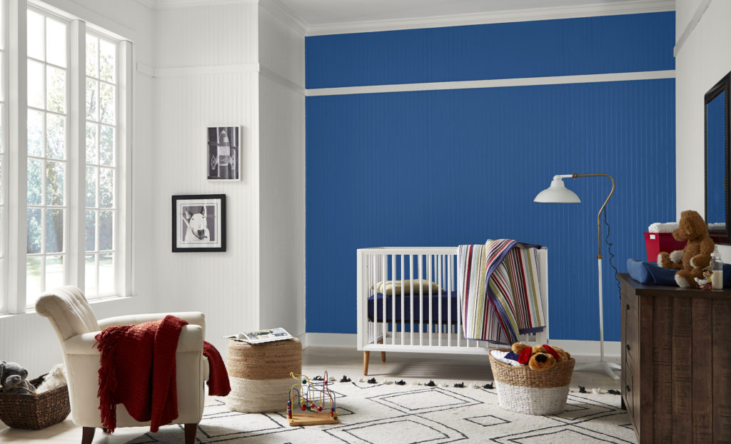 A colorful and casual nursery.  The room has a large window with an open garden vista.  The crib is white and modern with natural wood-tone legs. the room also features an accent walls with skinny shiplap painted in a blue color called Dark Cobalt Blue. 
