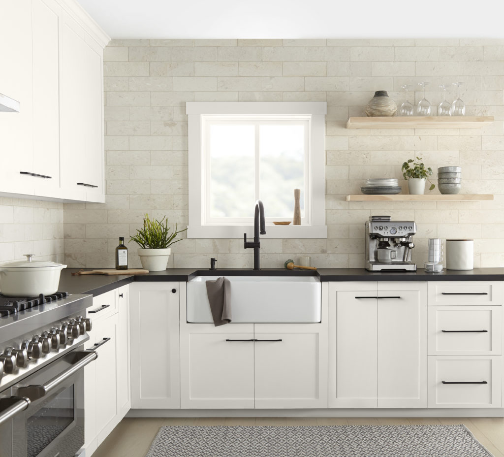 A modern farmhouse kitchen featuring white cabinetry painted with a warm white.  The countertop is dark granite and the backsplash is cream marble. The everyday essential servinignware can be on display on two natural wood free standing shelves. 