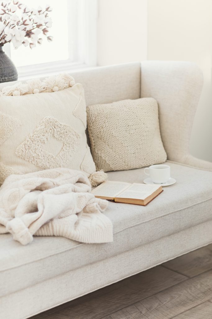 Still life details in the living room home interior. Woollen sweater, book and a cup of coffee on beige couch with knit macrame cushions. Cozy autumn or winter concept. Hygge style. The walls are painted with Blank Canvas.