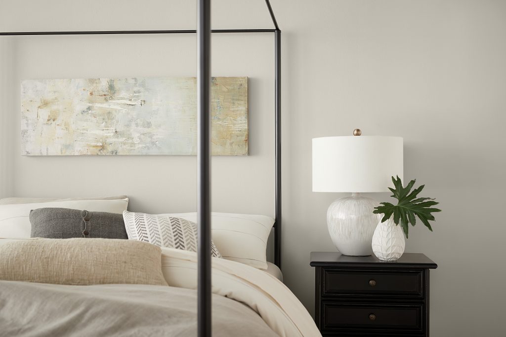 A modern casual bedroom, the color on the walls is a delicate taupe-gray called Tranquil Gray. The black frame canopy bed is dressed with layered neutral bedding and pillows.   