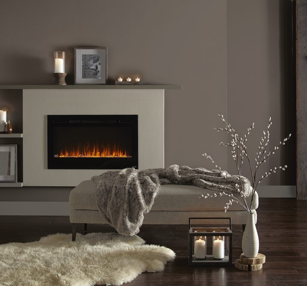 The corner of a contemporary living room features a lit fireplace, a comfy bench, a blanket and candles. Kindling is a dusty brown color used on the walls. 