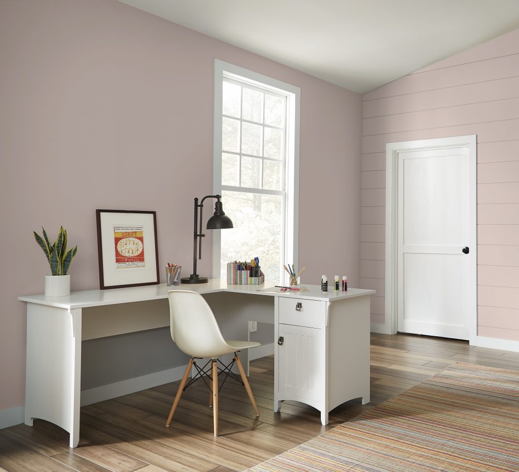 A home office / craft room that includes a work space with a white desk and a modern chair. A window with an outdoor vista and natural light coming into the room.  The room is painted with a pink paint color. 