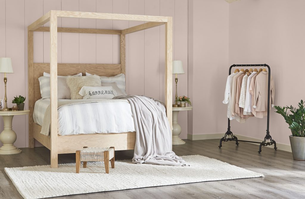 A large bedroom with a natural light wood canopy bed.  There is a garment rack with white and pink blouses.  