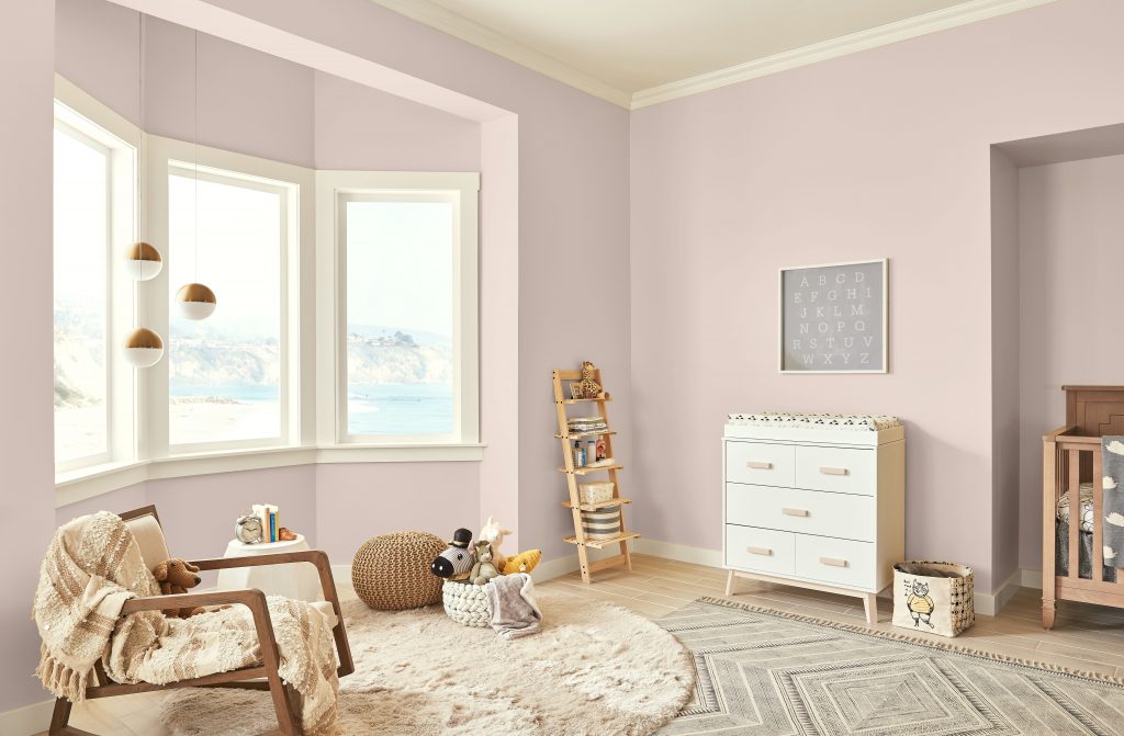 A pink kids room, there are several pieces of furniture that are made of natural wood.  Neutral toned carpet and blankets complete the pink room.  The is a bay window overlooking the beach and ocean water. 