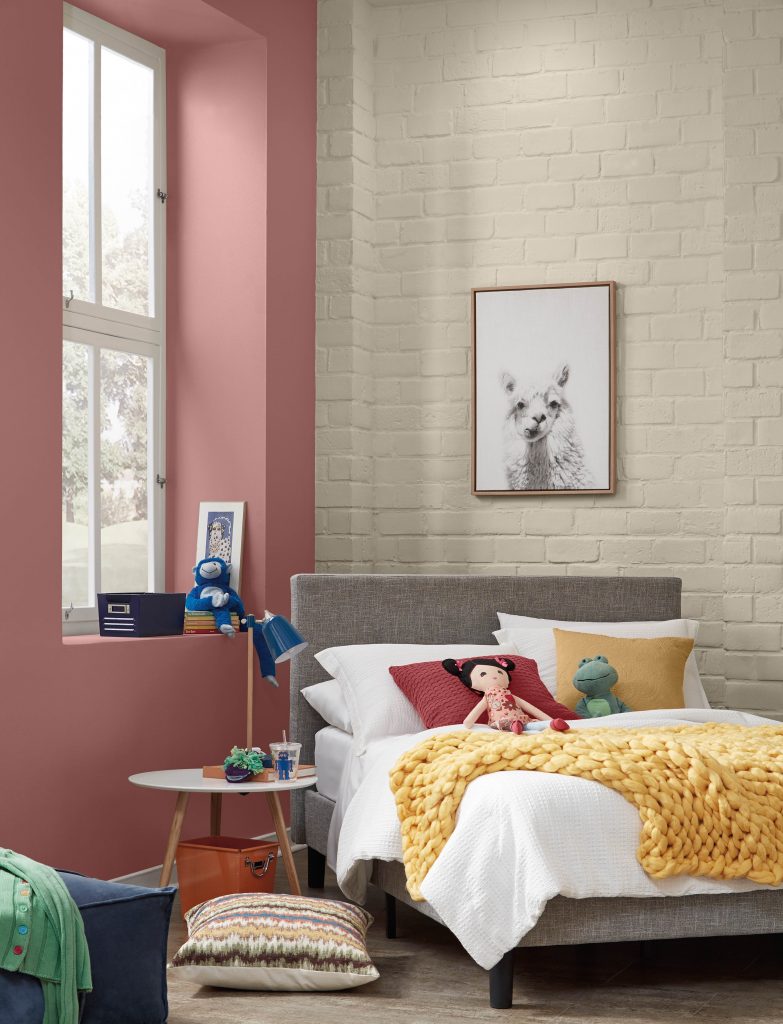 The corner of a kid's bedroom-playroom area.  There is a neutral brick wall featuring a beige color.  The accent wall on the left is a light red called Vermilion.