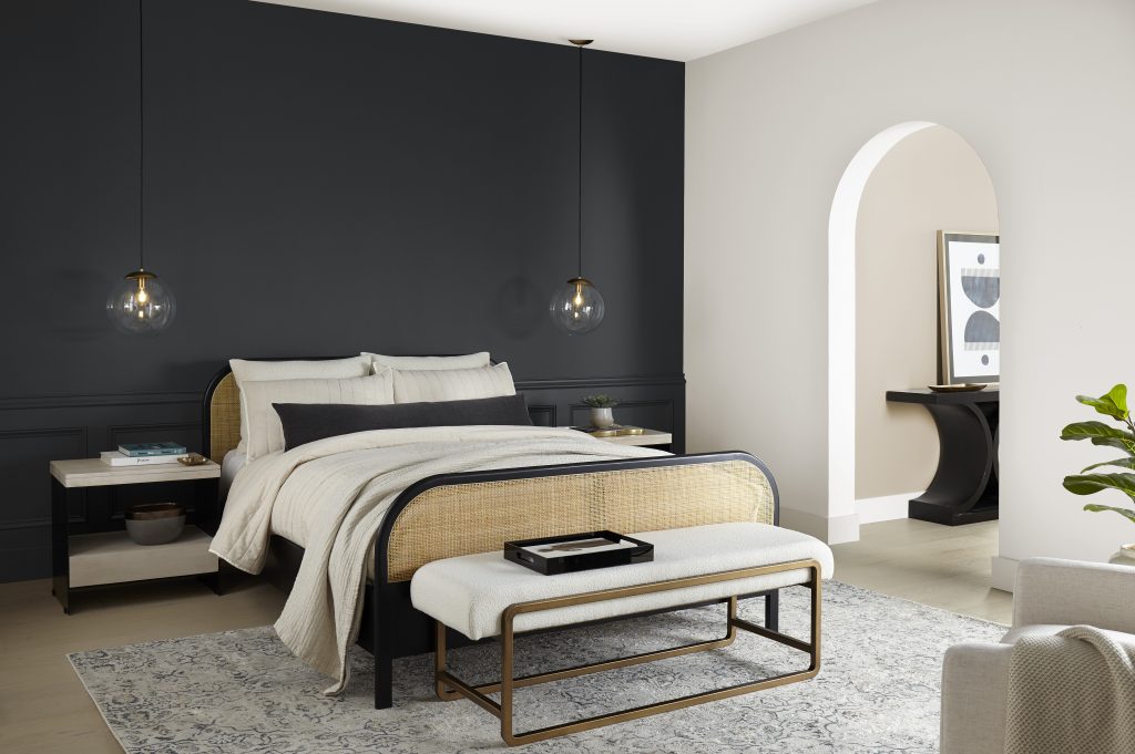 A large stylish bedroom with some art deco revival elements such a cane bed with a black frame and a soft modern bench at the end of the bed. 
 The accent wall behind the bed's headboard is painted in a sophisticated soft black called Cracked Pepper. There is an arched doorway that looks into the next room. 