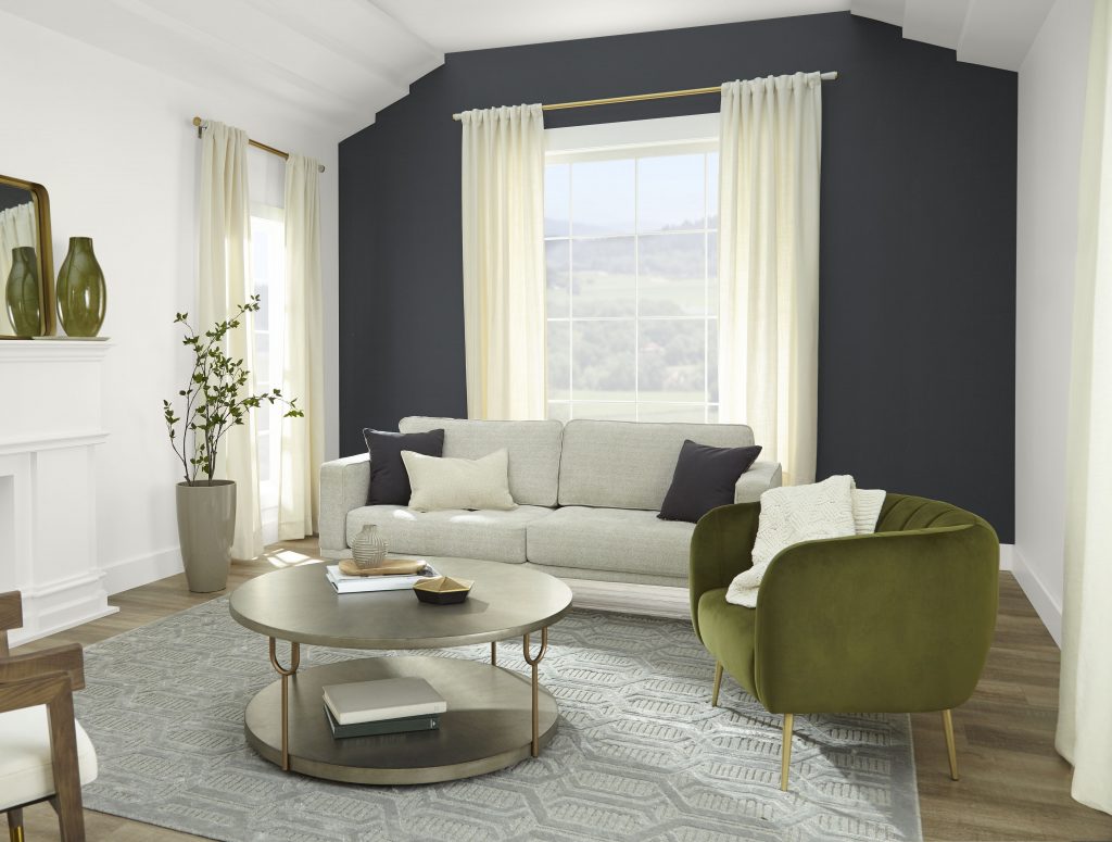 An Art Deco revival living room featuring a fireplace and an accent wall using BEHR's 2024 color of the year, Cracked Pepper.  There is a round coffee table, a green velvet accent chair, a light neutral modern sofa and the windows have floor length drapery. 