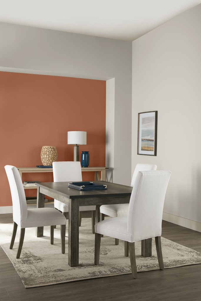 A casual dining room with simple furniture: a rectangular wooden table and simple chair with white color upholstery. Most walls are paint in a neutral color called Tranquil Gray and there is an accent wall painted with Orange Flambe. 