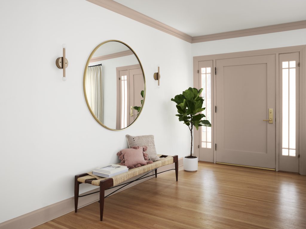 An entryway with a bench and a large round mirror.  The walls are white, and the baseboard, crown molding, trim, and front door are painted rosy taupe. 