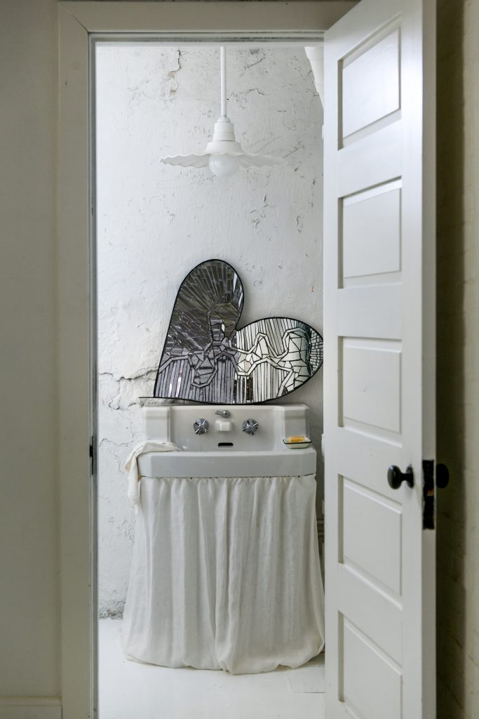 A bathroom with all white wall and a mirrored heart shape leaning above the sink.