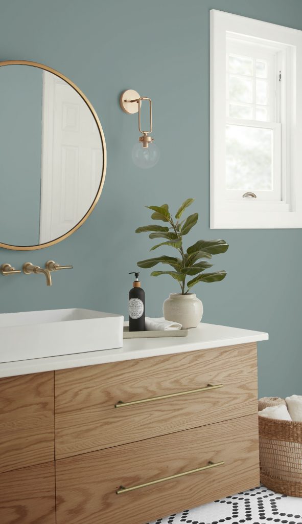 A Scandinavian style bathroom featuring a bold black and white pattern on the floor tile.  The vanity is a medium warm wood tone with with a large wood grain and the hardware is a metallic gold tone.  The walls are painted in cool tone called Provence Blue. 