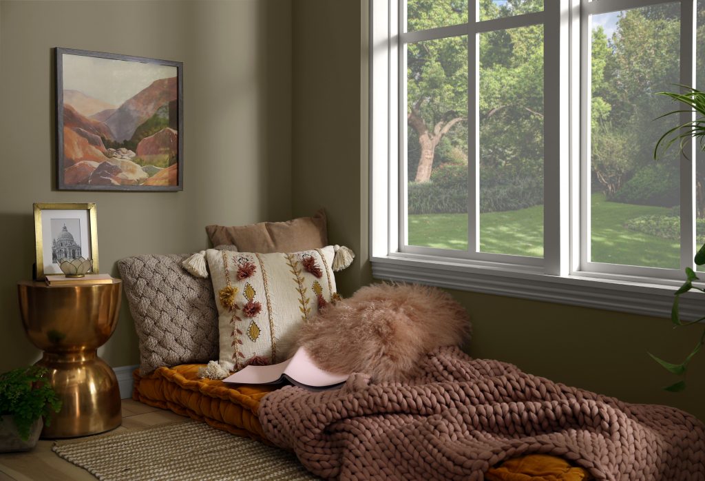 A quiet cozy are with blankets and pillow sitting next to a window. The walls are painted in an olive green hue.