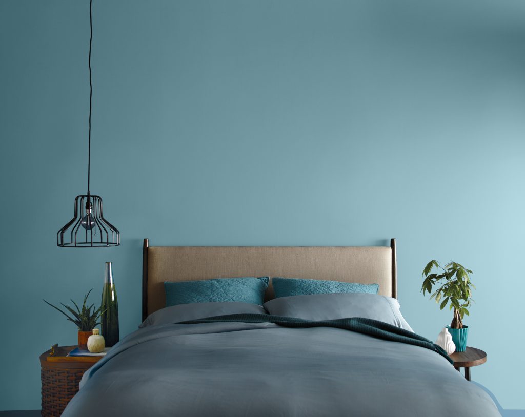 One wall of a bedroom with wall painted in Voyage. 
Brown headboard with blue furnishings on bed. Two side tables with plants and decorative elements.